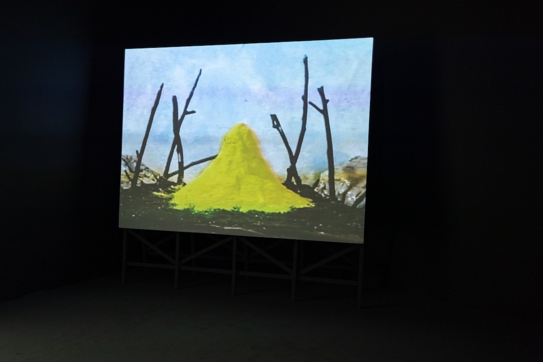 A towering projection screen with a wooden scaffold for a base appears in a dark theater. On the screen, a pyramid of bright yellow, sulphurous powder sits atop dark soil in the middle of a model landscape. Powder accumulates around the base of the heap, spreading out towards several blackened, spindly, upright twigs that resemble dead trees. Grey rocks appear on the outer edges of the makeshift horizon, pressed up against a mottled sky painted in blue and white.