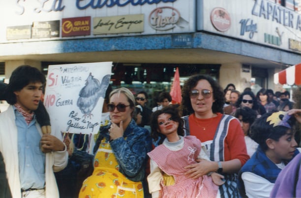 The scene of a street protest with two women—Maris Bustamante and Mónica Mayer—in the foreground and a large crowd behind them. Bustamante holds a sign that reads: "Si a la maternidad voluntaria / No a la guerra," while Mayer holds a doll. Both artists wear aprons with Styrofoam bellies.