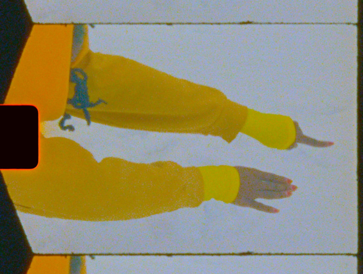 A performer reaches beyond the frame of the image in the midst of a choreographic act of tracing their surroundings. They wear layers of yellow, red nail polish, and a small trail of their hair completes their silhouette.