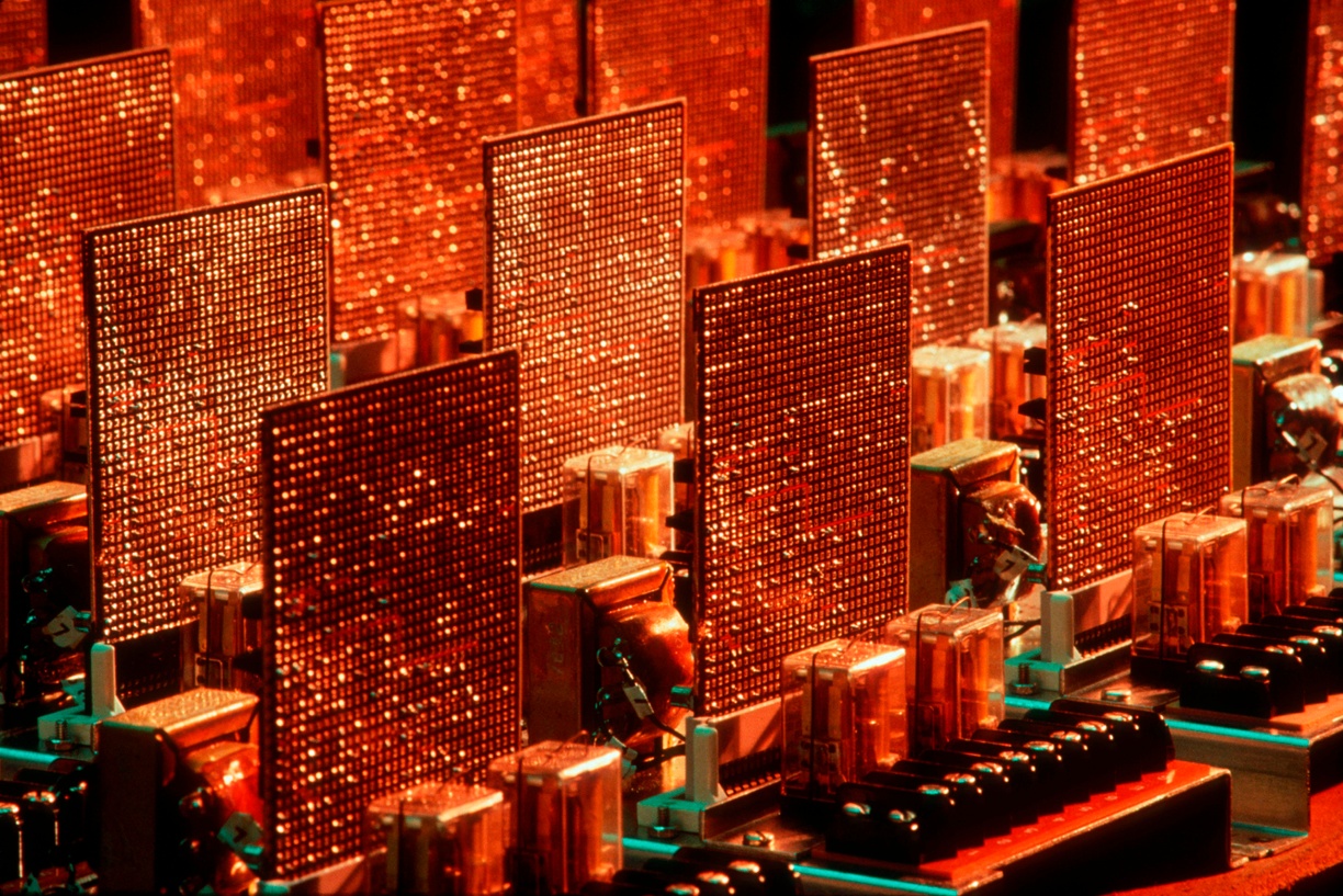 Taken by the photojournalist Erich Hartmann in 1982, this vibrant, color photograph features a close-up of an IBM circuit board for a computer. Taken at a slight angle, Hartmann shows three rows of three power amplifiers, with the fourth row cropped and pictured nearly out of frame. They appear as if they are rectangular-shaped skyscrapers, with lights dazzling across thousands of windows. The power amplifiers in the foreground are crisp and in focus, and as the image recedes, they become slightly blurred. The overall hue of the photograph is a burnt orange color, with a few turquoise highlights streaked across the body of specific hardware parts caused from the camera’s flash.