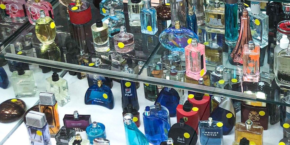 The inside of a store vitrine, dozens of multi-colored glass perfume bottles sit on two shelves. Many bottles have a round bright yellow sticker placed on them.
