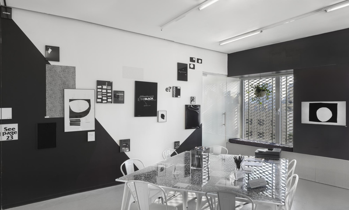 Installation photograph documenting a room within the art exhibition *The Black School X Kameelah Janan Rasheed* organized at the New Museum in New York. In the center of the room is a table for workshops and handling archival materials. Behind the table on the walls are various wall-mounted text and abstract artworks by artist Kameelah Janan Rasheed.